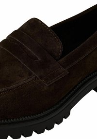 Bruna mockaläder loafers med ett upphöjt remdetalj. Sulan är texturerad och robust, vilket kontrasterar med det mjuka ovandelmaterialet.