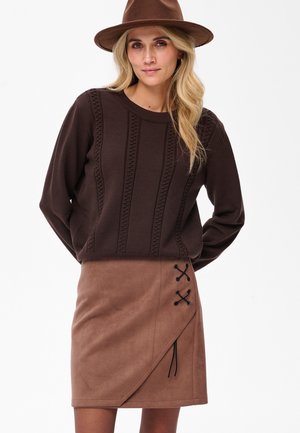 Brauner gestrickter Pullover mit strukturierten vertikalen Streifen, kombiniert mit einem braunen Wildlederimitat-Rock, der mit einer seitlichen Schnürung und asymmetrischem Saum versehen ist.