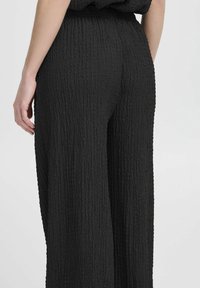 Schwarze, weit geschnittene Hosen aus strukturiertem Stoff mit einer zerknitterten Optik. Verfügt über einen elastischen Bund für optimalen Tragekomfort.