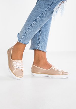 Frau trägt hellblaue zerrissene Jeans und glänzende beige Schnürschuhe, steht auf weißem Boden.
