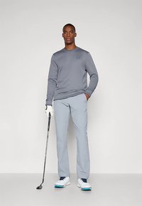 Hombre con camisa de manga larga gris y pantalones gris claros sostiene un palo de golf, mientras lleva un guante blanco y zapatos de golf blancos.