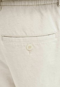 Gros plan de la poche arrière beige d'un pantalon avec une fente horizontale et un petit bouton beige sous l'ouverture de la poche.