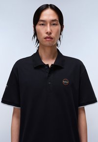 Polo shirt nera con vestibilità rilassata, dotata di una chiusura a tre bottoni e di un logo circolare sul petto. Dettagli grigi sul colletto.