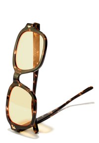 Ochelari de soare tortoiseshell de culoare maronie, cu lentile pătrate supradimensionate și sticlă colorată în galben. Au brațe elegante și o marcă subtilă pe ramă.