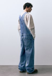 Lichtblauwe denim tuinbroek met een losse pasvorm, verstelbare bandjes en zijzakken. Draagt over een beige T-shirt met print en bruine laarzen.