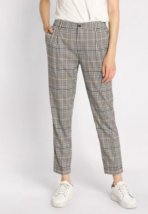 Personne portant un pantalon ajusté taille haute à carreaux noir, blanc et beige, associé à des baskets blanches et un haut blanc rentré.