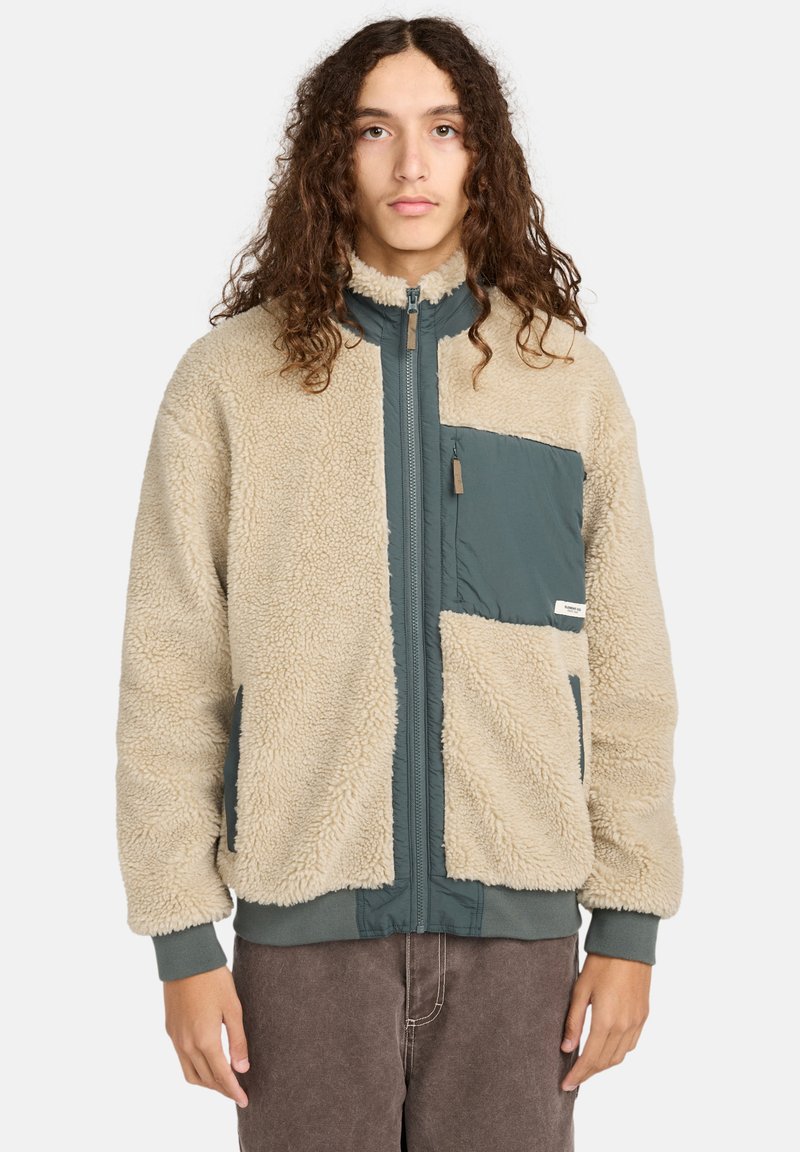 Flauschige beigefarbene Jacke mit tealfarbenem Panele und Brusttasche. Merkmale hoher Kragen, Reißverschluss und gerippte Bündchen. Strukturierter Stoff insgesamt.