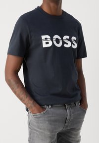 En marinblå bomulls-T-shirt med en djärv, vit "BOSS"-logotyp, rund halsringning och korta  ärmar. Bärs tillsammans med grå jeans och synlig arm-tatuering.
