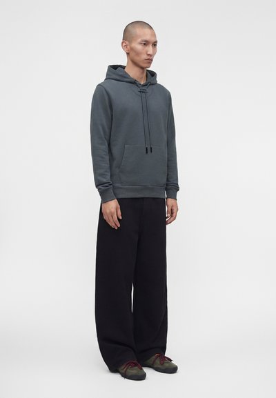 Homme debout portant un sweat à capuche gris foncé avec poche avant, pantalon noir ample et baskets vertes avec lacets rouges sur un fond clair uni.