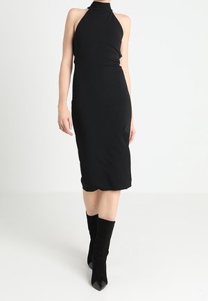 Vrouw die een zwarte midi-jurk zonder mouwen draagt met een hoge halslijn en zwarte enkellaarsjes met puntige neus, staand op een witte achtergrond.