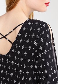 Top de tela negra con un patrón geométrico blanco, con un escote bajo y un diseño de tirantes entrecruzados. Material ligero y texturizado.