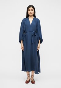 Γυναίκα που στέκεται φορώντας μπλε φόρεμα μέτριου μήκους με μακριά μανίκια, ντεκολτέ V, δεμένη στη μέση και με πλευρικά ανοίγματα, με καφέ δερμάτινα παπούτσια με κορδόνια και μεγάλο σκουλαρίκι.