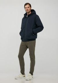 Giacca con zip blu navy e cappuccio, dotata di due tasche laterali e polsini a costine, abbinata a pantaloni verde oliva a gamba affusolata e sneakers bianche.