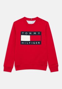 Crvena majica s okruglim izrezom, s velikim crnim logotipom u obliku kutije s tekstom "TOMMY HILFIGER" i ukrasnim obojenim pravokutnicima. Rebrasti manšeti.