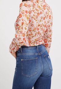 Floral gemusterte, halbtransparente Bluse in Hellrosa mit orangen Akzenten, kombiniert mit hoch taillierten, dunkelblauen Denim-Jeans mit Gesäßtaschen.