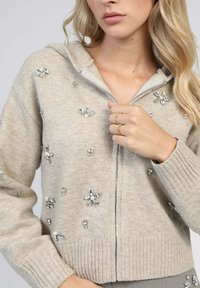 Sweat à fermeture éclair beige fabriqué en tissu doux, avec des embellissements floraux et des accents argentés. Poignets et ourlet côtelés pour une texture supplémentaire.