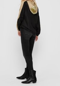 Schwarzer Windbreaker mit einer beigen und reflektierenden Kapuze, kombiniert mit dunklen Skinny-Jeans und schwarzen Stiefeletten, von hinten gesehen.