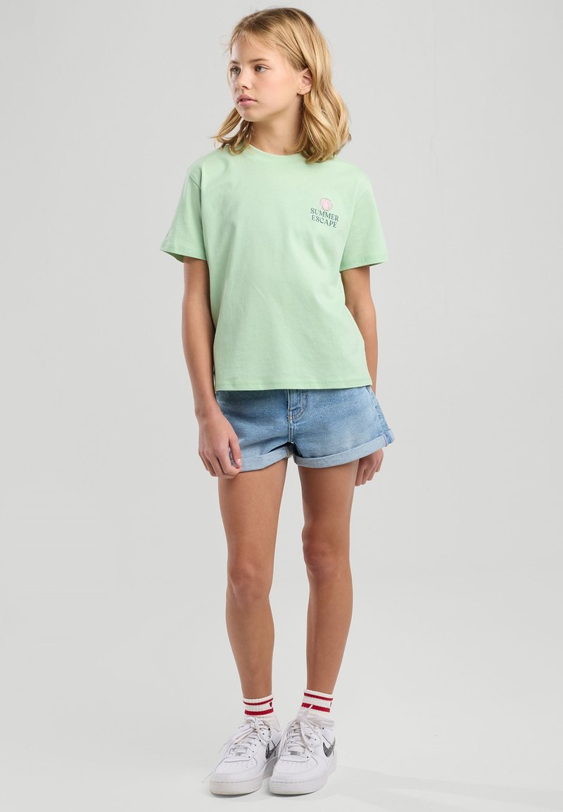 Mint green cotton t-shirt with “SUMMER ESCAPE” printed on the chest, paired with light blue denim shorts. White sneakers and red-striped socks.