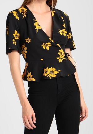Frau trägt eine schwarze Wickelbluse mit gelbem Blumenmuster und kurzen Ärmeln, kombiniert mit hoch taillierter schwarzer Hose.