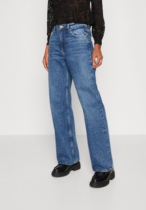 Personne portant un jean bleu coupe droite, des chaussures noires épaisses et un haut en dentelle noire à manches longues sur un fond blanc uni.