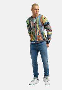 Mønstret striktrøje i mange farver med abstrakte mønstre, parret med blå jeans og hvide sneakers, der har grønne detaljer og opkrøllede ærmer.