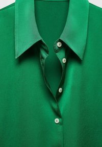 Chemise en satin vert avec un col pointu, une patte de boutonnage à cinq boutons ronds blancs, et une texture lisse et brillante.