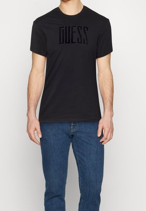 Camiseta de algodón negra de manga corta con un logotipo de textura de terciopelo "GUESS" colocado de manera prominente en la parte frontal. Combinada con jeans azules.