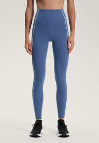 Visoke modre legice iz raztegljive tkanine z belimi stranskimi šivi, prilegajoča zasnova in gladka tekstura, v kombinaciji s črnimi športnimi copati.