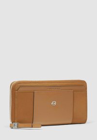 Portafoglio con cerniera in pelle color cuoio, con tasca frontale, logo argentato e cinturino da polso su sfondo grigio chiaro.