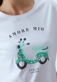 Vit bomullströja med en grafik av en turkos scooter med gröna paljetter. Texten lyder "AMORE MIO ITALY... ITALIENSKA SOMRAR."