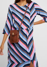 Donna che indossa un vestito a righe diagonali blu, rosa, marrone e nero, mentre porta una piccola borsa a tracolla marrone con una chiusura dorata.