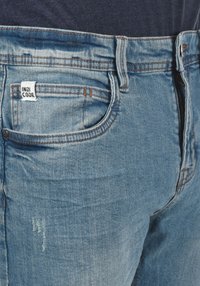Falmede blå denim jeans med en frontlomme, lynlåsclosure og et synligt mærke, der læser "INDI CODE." Lyse slidte detaljer på stoffet.