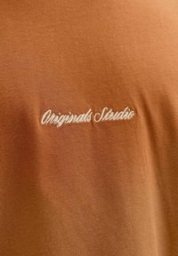 Smeđa majica s kratkim rukavima izrađena od mekog materijala, s izvezenim tekstom "Originals Studio" u bijeloj boji. Jednostavan, neukrašeni dizajn.