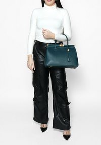 Teal leren handtas met een gestructureerde vorm, gouden hardware en een bovenhandvat. Het lijkt textuur te hebben en beschikt over een klein hanglabel.