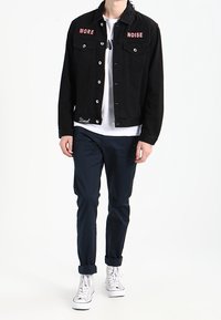 Černá džínová bunda s červeným vyšívaným textem "MORE" a "NOISE", dvě kapsy na prsou a knoflíky. Kombinováno s tmavě modrými kalhotami a bílými teniskami.
