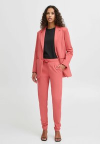 Korálově růžový blazer a odpovídající tepláky, vyrobené z hladké tkaniny. Sako má dvě přední kapsy a kalhoty mají elastický stahovací pas.