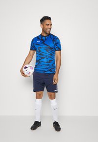 Blau- und schwarz gemustertes Sporttrikot, kombiniert mit marineblauen Shorts, weißen Socken und schwarzen Fußballschuhen; der Spieler hält einen Fußball.