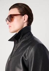 Black leather jacket with a high collar, featuring a zip closure. Model wears oversized brown sunglasses, highlighting a smooth, sleek texture.
