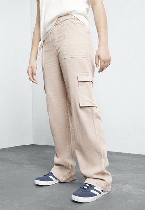 Hellbeige Cargohosen mit seitlichen Taschen, geradem Bein und elastischem Bund, kombiniert mit marineblauen und weißen Sneakers.