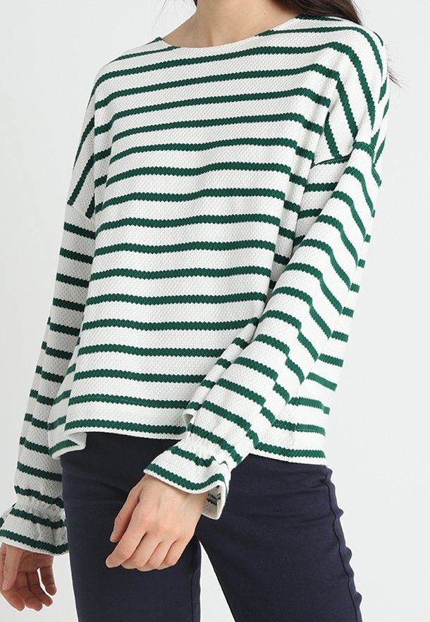 Nainen, jolla on päällään valkoinen pitkähiha usein vihreillä vaakaraitoja ja röyhelöhihoja, yhdistettynä tummiin housuihin.