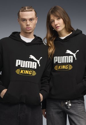 Jonge man en vrouw dragen zwarte Puma King hoodies en staan naast elkaar tegen een grijze achtergrond.