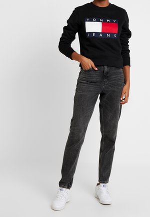 Persona che indossa una felpa nera Tommy Jeans, jeans grigio scuro e sneakers bianche, in piedi con una mano in tasca su sfondo grigio.