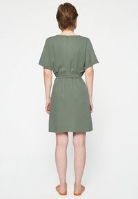 Robe verte à manches courtes avec une taille cintrée, un ourlet arrivant au genou et un tissu lisse. Sandales plates portées pieds nus.