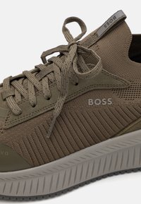 Shoe atlético castanho com parte superior de tecido texturizado, detalhe em malha, atacadores planos e uma sola cinza larga e estriada. O logótipo está visível de lado.