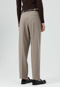 Pantalon beige cintré avec une coupe ample, doté d'une poche arrière unique, assorti à des chaussures noires. Le matériau semble lisse avec un léger éclat.