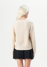 Beige gebreide trui met een gestructureerd patroon, lange mouwen en een ronde halslijn. Gragen over een zwarte plooienrok.