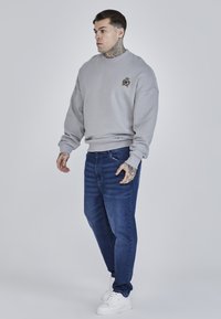 Hanorac gri deschis cu guler rotund și detalii cu emblema aurie, purtat cu blugi bleu închis și adidași albi. Textură netedă pe toată suprafața.