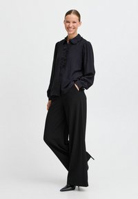Camicia nera con frange sul davanti, chiusura a bottoni e colletto, abbinata a pantaloni neri ampi e tacchi a punta neri, che mostrano una silhouette elegante.