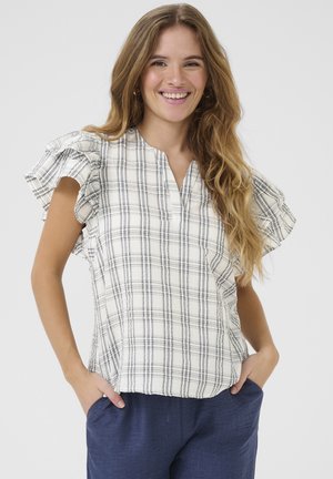 Ung kvinde smilende, iført en hvid ternet kortærmet bluse med flæsedetaljer og mørkeblå bukser, hænderne i lommerne.