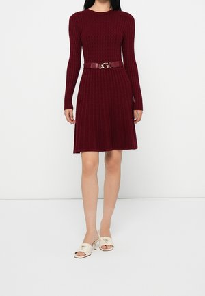 Femme portant une robe bordeaux en maille torsadée à manches longues avec une ceinture assortie et des sandales blanches à talons ouverts, debout devant un fond blanc.
